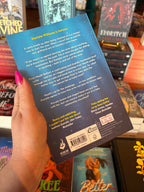 A hand with pink glitter nail polish holds the back of the book 'Better Than Revenge' by E.J. Blaise, featuring an underwater scene with yellow and white font for the blurb. The book is held up in a bookshop surrounded by other blurry books.