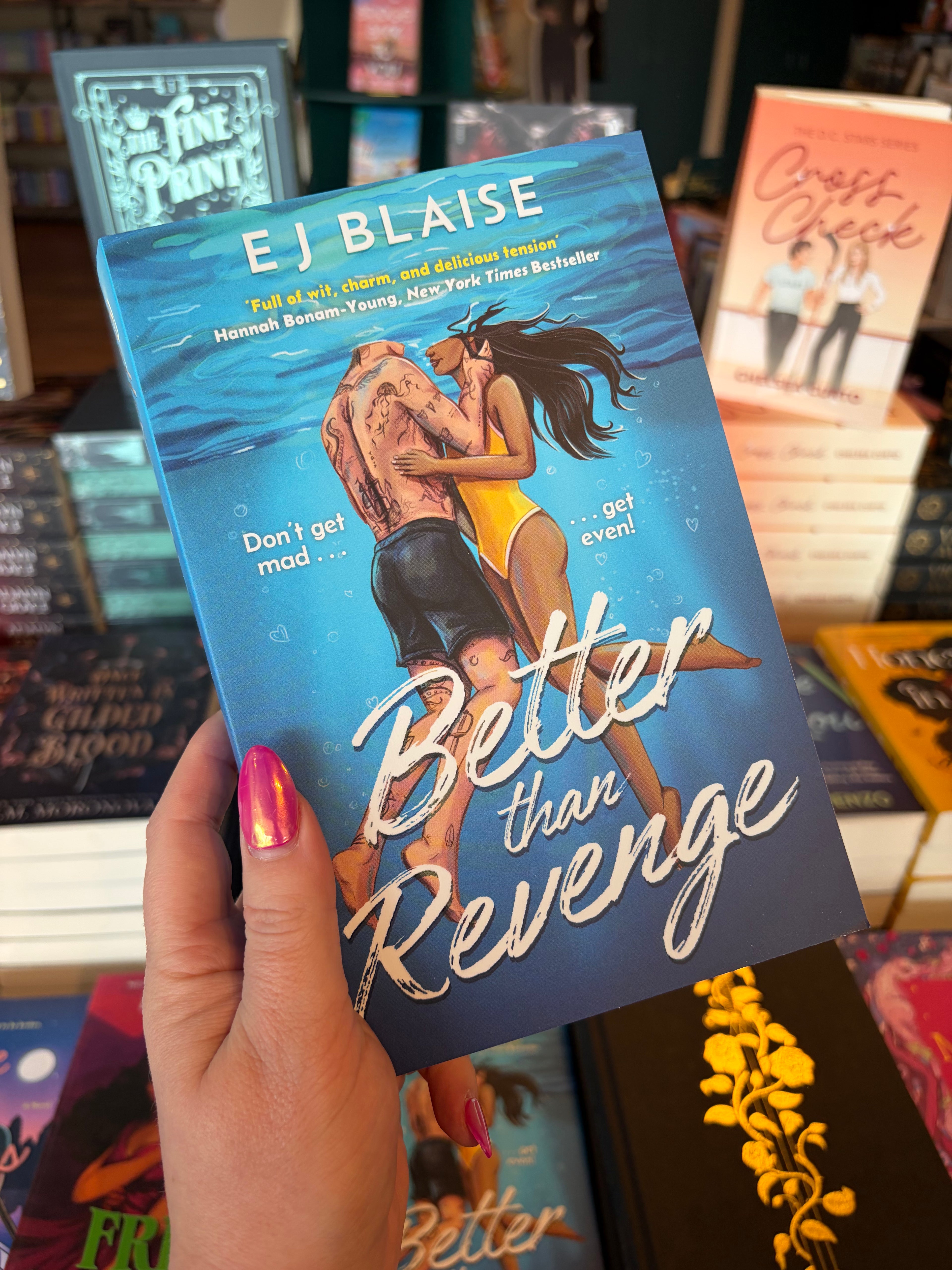 A hand with pink glitter nail polish holds a copy of the book 'Better Than Revenge' by E.J. Blaise, featuring an illustration of a couple embracing underwater. He is wearing black swimming trunks and covered in tattoos, she has a yellow one piece glowing against her dark skin. The book is held up in a bookshop surrounded by other blurry books.