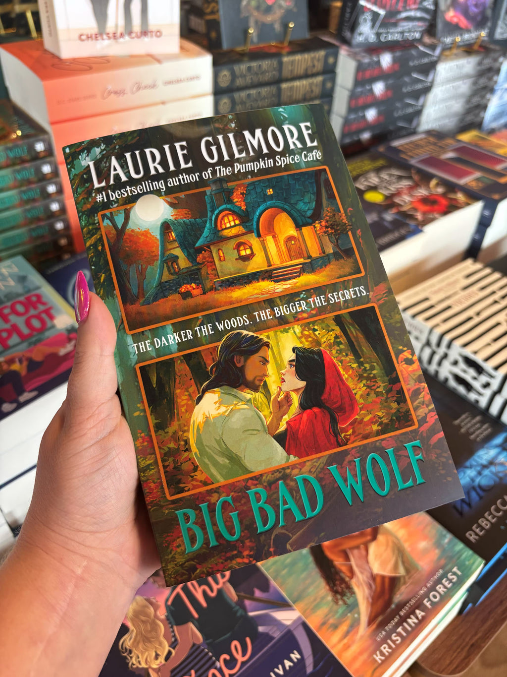 A hand with pink nail polish holds a copy of 'Big Bad Wolf' by Laurie Gilmore in a bookstore with blurry books in the background. The cover features a forest scene and two gold comic-style rectangles, one with a cottage and the other with a man and a woman in an embrace frowning at each other. 