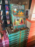 A stack of the book 'Big Bad Wolf' by Laurie Gilmore set in a bookstore, with a cover featuring a forest scene and two gold comic-style rectangles, one with a cottage and the other with a man and a woman in an embrace frowning at each other. 