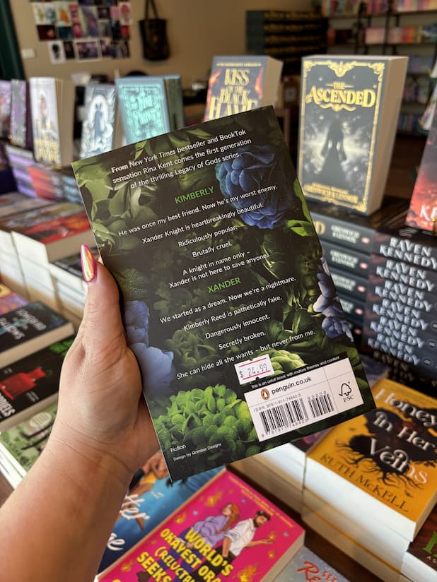 A hand with pink nail polish holds the back of a copy of 'Black Knight' by Rina Kent, featuring a green and blue floral design with white text.