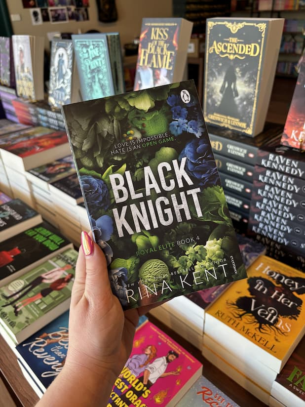 A hand with pink nail polish holds a copy of 'Black Knight' by Rina Kent, featuring a green and blue floral design with large white text.