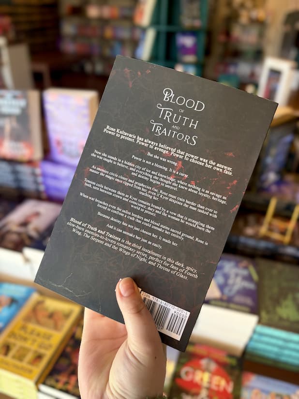 A hand holding the back of a copy of 'Blood of Truth and Traitor' by Aria Ashbrook, featuring a dark ornate background with white text.