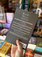 A hand holding the back of a copy of 'Blood of Truth and Traitor' by Aria Ashbrook, featuring a dark ornate background with white text.