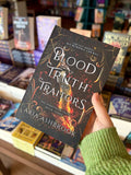 A hand holding a copy of 'Blood of Truth and Traitor' by Aria Ashbrook, featuring a dark ornate background with a large sword and glowing fire, plus white title text and author name.