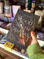 A hand holding a copy of 'Blood of Truth and Traitor' by Aria Ashbrook, featuring a dark ornate background with a large sword and glowing fire, plus white title text and author name.