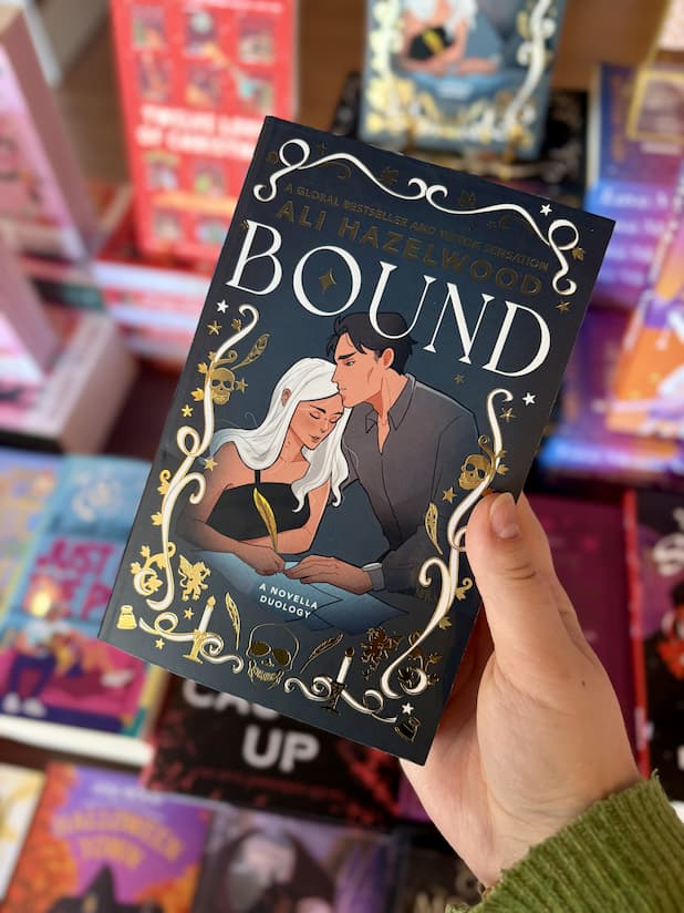 A hand holds the book 'Bound' by Ali Hazelwood. The book cover is dark navy blue with gold foil details, white gothic lettering, and an illustration of a man and woman embracing.