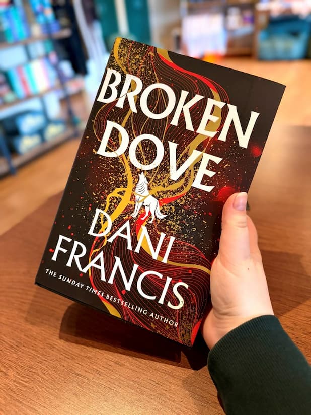 A hand holding a copy of the hardcover edition of 'Broken Dove' by Dani Francis, featuring a white wolf in between large, white text and gold and red detailing.