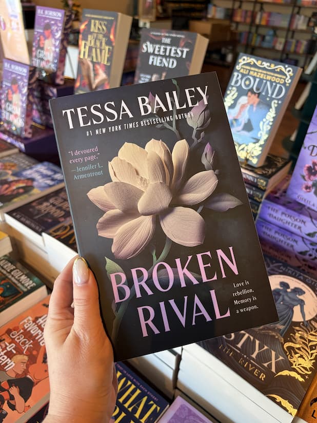 A hand with nude nail polish holding a copy of 'Broken Rival' by Tessa Bailey, featuring a white flower on a dark background, and large text.