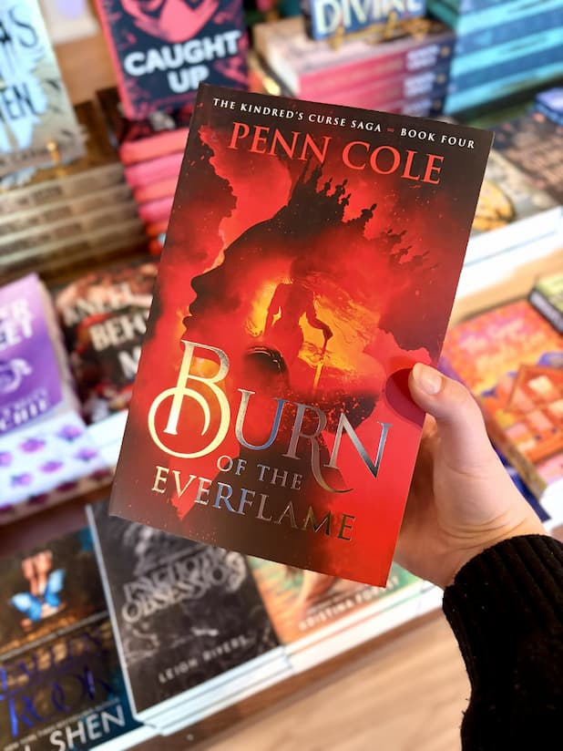 A hand holding a copy of 'Burn of the Everflame' by Penn Cole, featuring a dramatic red and black fiery illustration with a silhouetted figure and large white title text.
