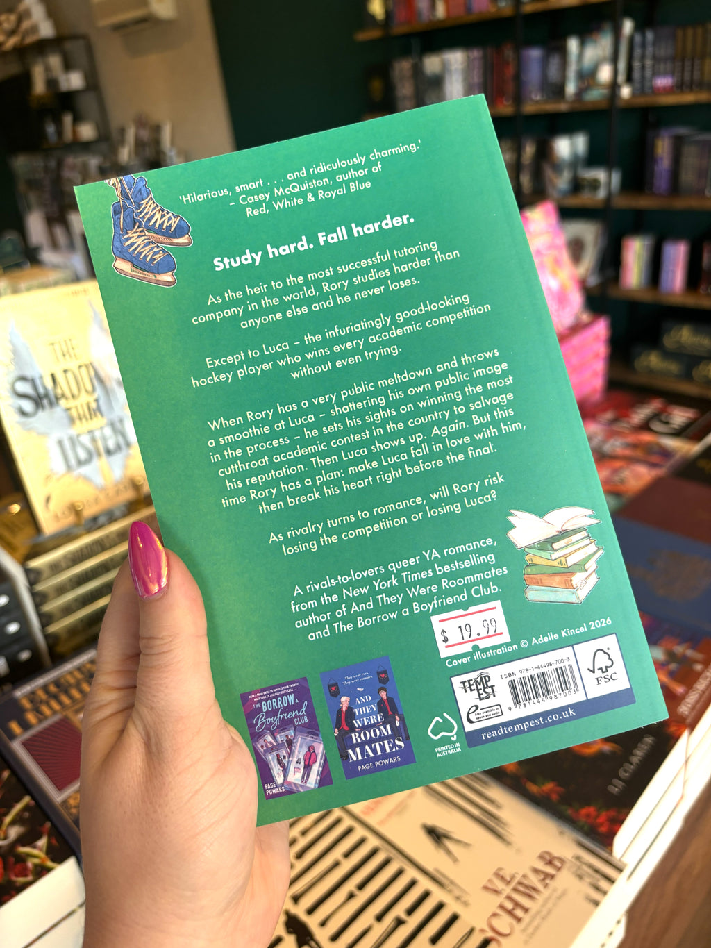 A hand with pink nail polish holding the back of a copy of 'But I Hate Him' by Page Powars with a green background, an illustration of ice skates and a stack of books, and white text.