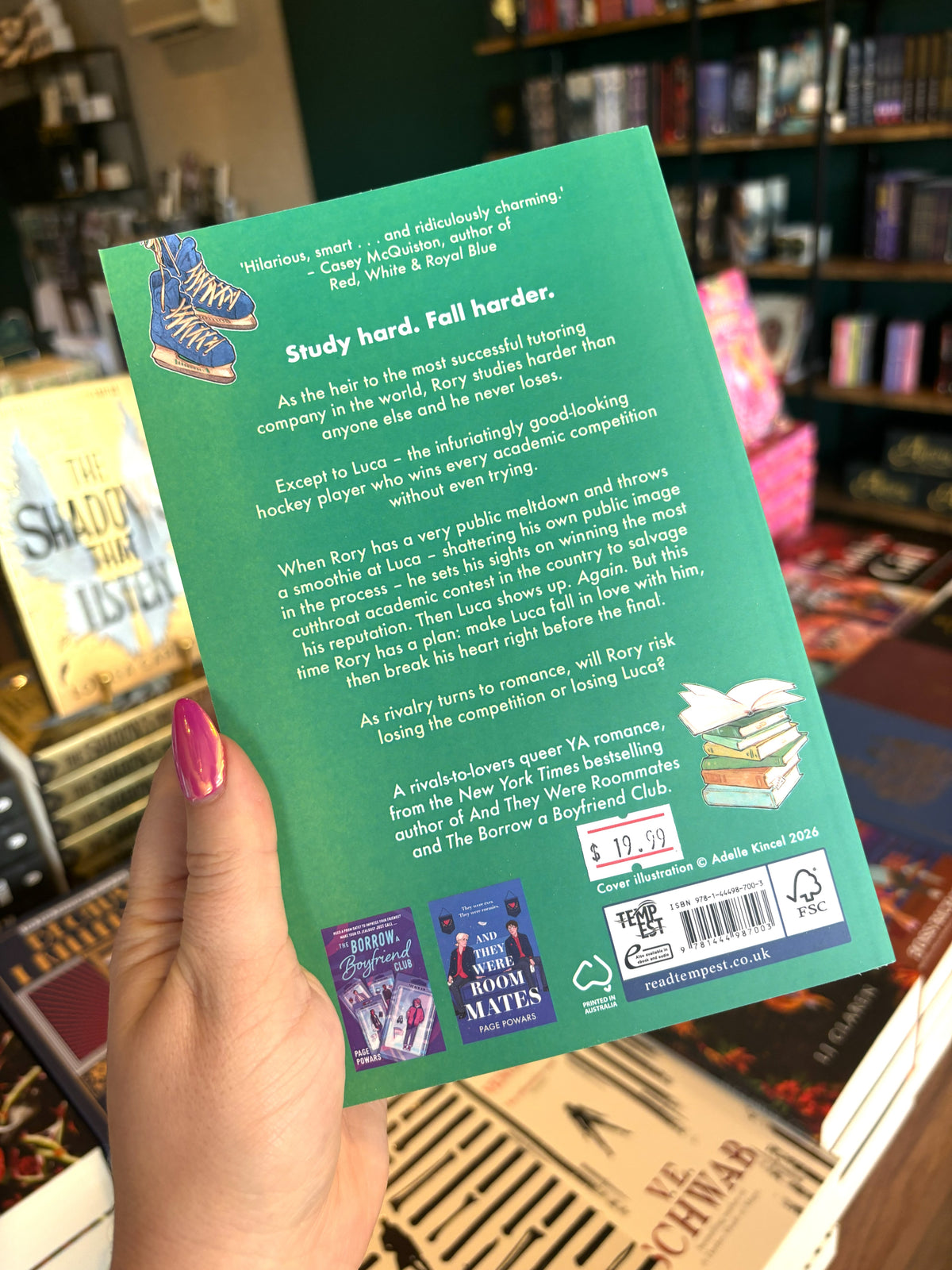 A hand with pink nail polish holding the back of a copy of 'But I Hate Him' by Page Powars with a green background, an illustration of ice skates and a stack of books, and white text.