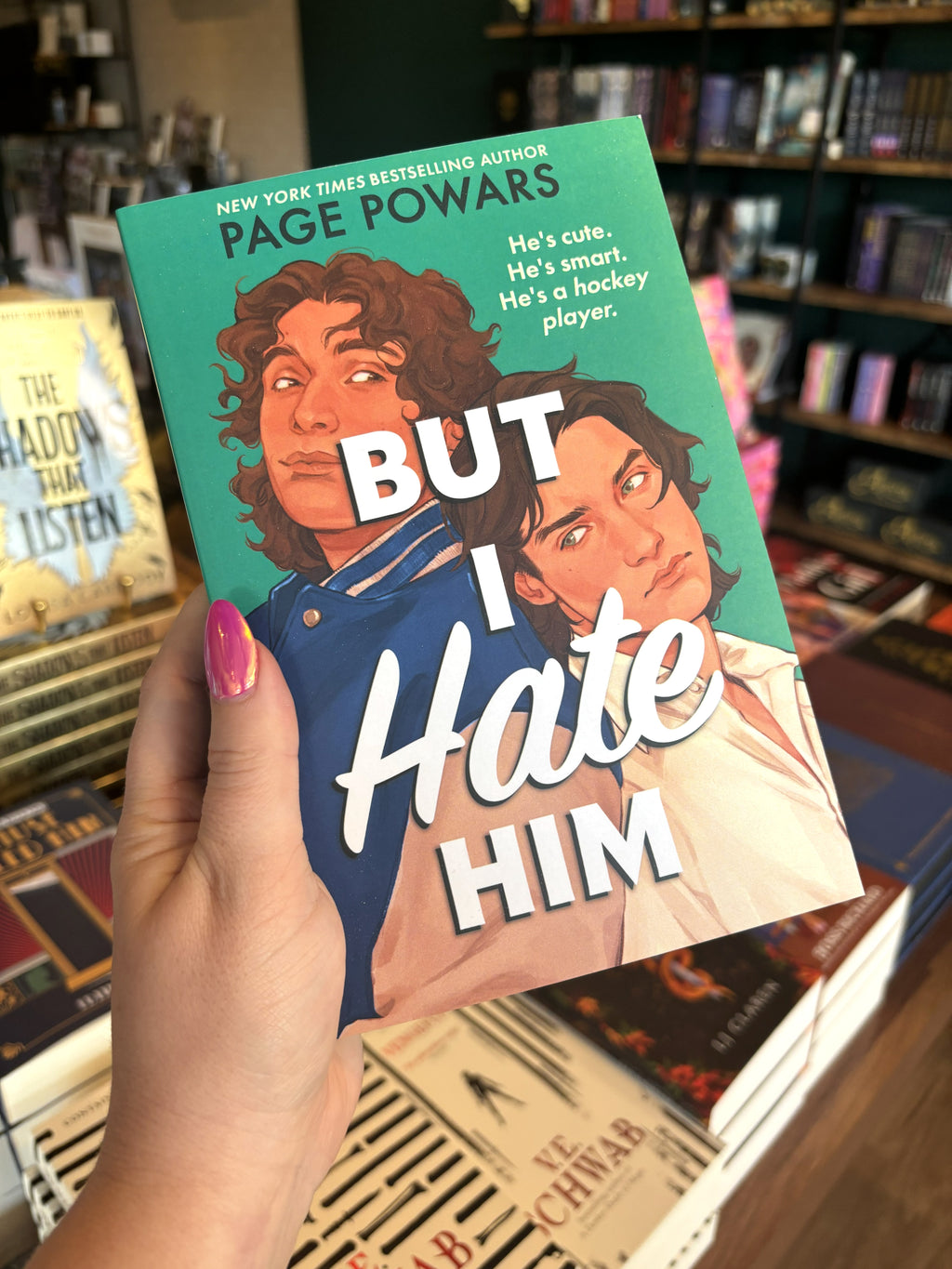 A hand with pink nail polish holding a copy of 'But I Hate Him' by Page Powars with two men back to back looking at each over their shoulders on a green background.