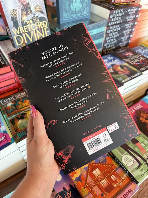A hand with pink nail polish holding the back of a hardcover copy of 'Caught Up' by Navessa Allen, featuring a black cover with jagged red shards of glass, and white text.