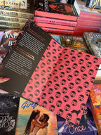 A hand with pink nail polish holding open a hardcover copy of 'Caught Up' by Navessa Allen, featuring red pages with red mask illustrations.