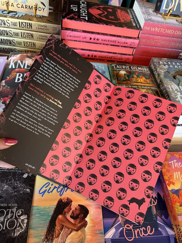 A hand with pink nail polish holding open a hardcover copy of 'Caught Up' by Navessa Allen, featuring red pages with red mask illustrations.