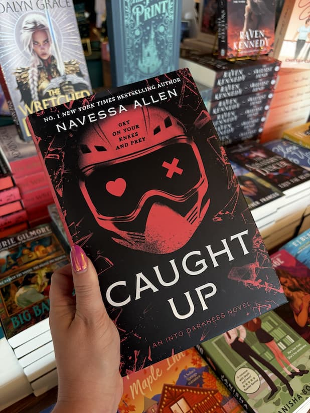 A hand with pink nail polish holding a hardcover copy of 'Caught Up' by Navessa Allen, featuring a red helmet design and red and white text on a dark background.
