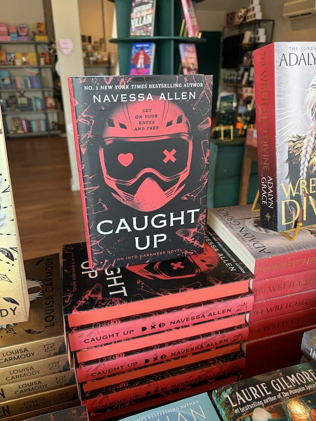A stack of hardcover copies of 'Caught Up' by Navessa Allen with a single copy standing on top, featuring a red helmet design and red and white text on a dark background.