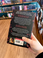 A hand holding the back of a paperback copy of 'Caught Up' by Navessa Allen, featuring a black cover with jagged red shards of glass, and white text.