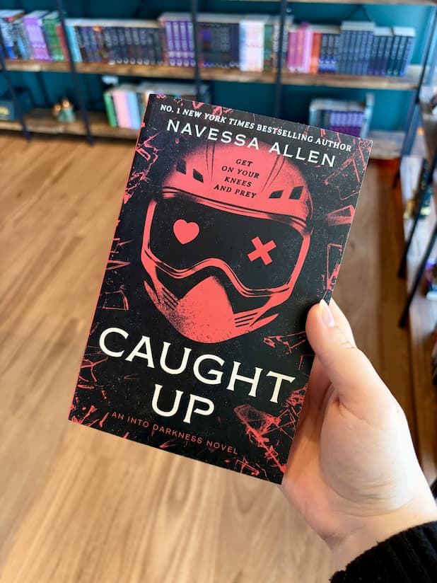A hand holding a paperback copy of 'Caught Up' by Navessa Allen, featuring a red helmet design and red and white text on a dark background.