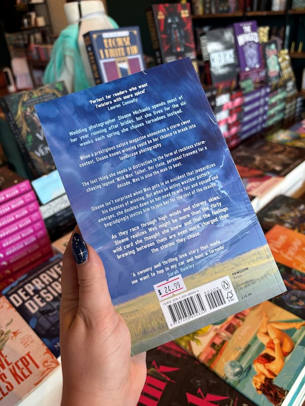 A hand with blue starry nail polish holds a copy of the book 'Chase Me if You Can' by Heather Frances, the back cover of the book blurb showcases the rivals to lovers storm chaser romance.
