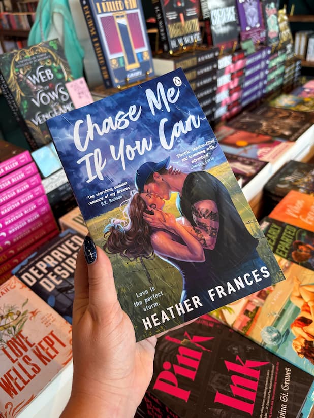 A hand with blue starry nail polish holds a copy of the book 'Chase Me if You Can' by Heather Frances, featuring a male/female couple embracing in a kiss in front of a stormy field. He is wearing a backwards cap and has tattoos down his left arm. 