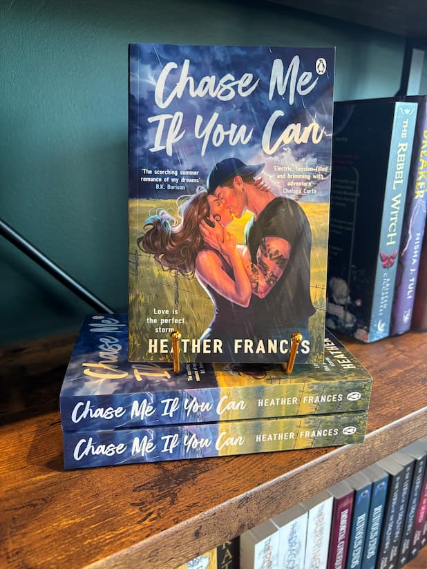 A book titled "Chase Me If You Can" by Heather Frances stands upright on a stack of two copies on a wooden bookshelf. The cover shows a blue stormy sky with a couple embracing in a kiss. The male wearing a backwards cap and tattoos down his left arm.