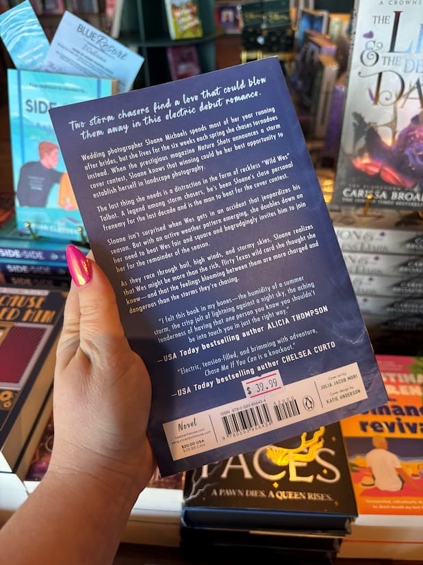 A hand holds the back cover of 'Chase Me if You Can' by Heather Frances, featuring a blue background with white font for the blurb. The book is held in front of a bookstore full of other blurry books in the background. 
