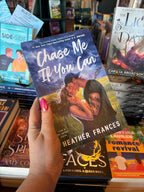 A hand holds the book cover of 'Chase Me if You Can' by Heather Frances, featuring a male/female couple embracing in a kiss in front of a stormy field. He is wearing a backwards cap and has tattoos down his left arm. The book in held in front of a table full of other blurry books.