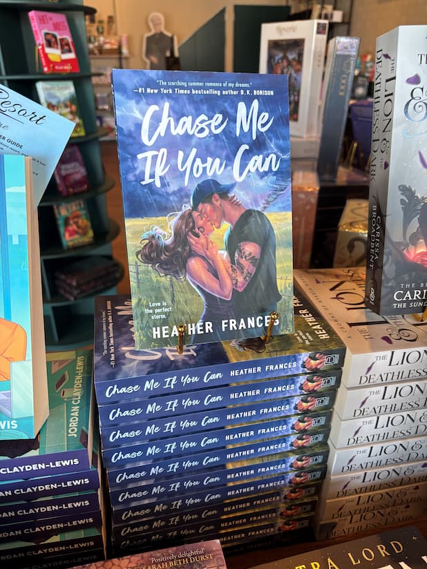 A stack of the book 'Chase Me if You Can' by Heather Frances, featuring a male/female couple embracing in a kiss in front of a stormy field. He is wearing a backwards cap and has tattoos down his left arm. The stack is located in a bookstore with other books blurry in the background.