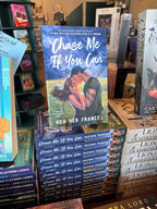 A stack of the book 'Chase Me if You Can' by Heather Frances, featuring a male/female couple embracing in a kiss in front of a stormy field. He is wearing a backwards cap and has tattoos down his left arm. The stack is located in a bookstore with other books blurry in the background.