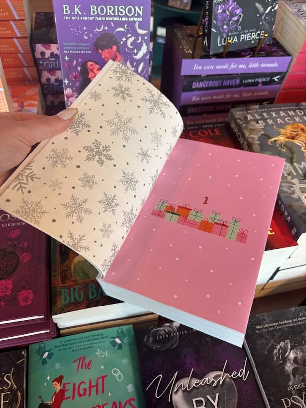 A hand with nude nail polish holds open a copy of the Christmas advent romance book 'Christmas Eve Love Story: An Advent Book' by Ginny Baird. The left page is white with grey snowflakes and the right page is pink with snow falling and a stack of presents under the number 1.