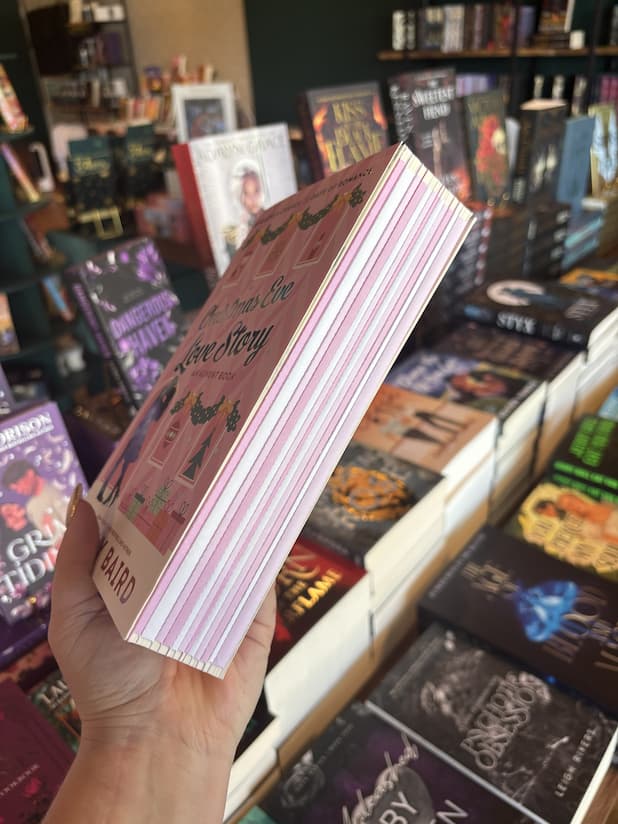 A hand with nude nail polish shows the side of a copy of the Christmas advent romance book 'Christmas Eve Love Story: An Advent Book' by Ginny Baird. The pages are wrapped in pink and white paper.