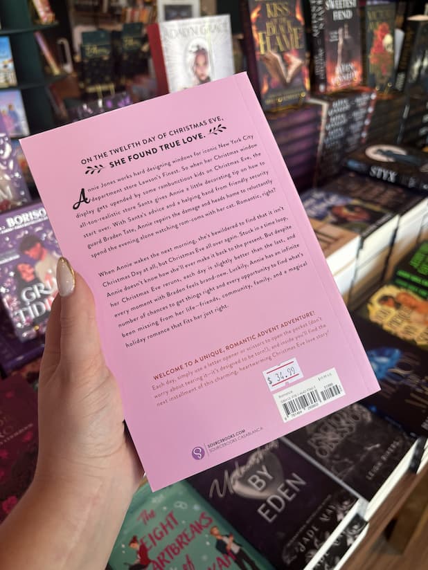 A hand with nude nail polish holds the back of a copy of the Christmas advent romance book 'Christmas Eve Love Story: An Advent Book' by Ginny Baird, featuring a pink cover with black text.
