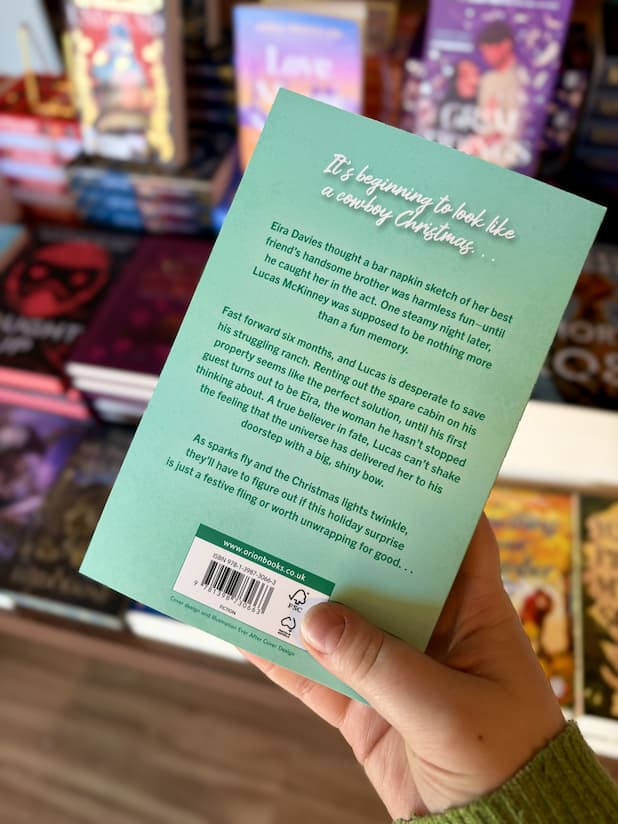 A hand holding the back of a copy of 'Christmas at Fox Ridge' by Bailey Hannah, featuring a light teal background with white and green text.