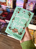 A hand holding a copy of 'Christmas at Fox Ridge' by Bailey Hannah, featuring a light teal background with white script title text, a decorative wreath border with holly, pinecones, ornaments, and a brown cowboy hat illustration.