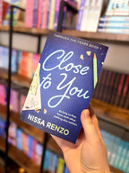 A hand holding the cover for 'Close to You' by Nissa Renzo, featuring a textured dark blue background with large light blue script title text, a green pencil, pencil shavings, and school-themed paper accents.