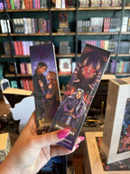 A hand with pink nail polish holds two physical book bookmarks featuring illustrated covers from 'Fae Lovers' and 'Dragon Princess' collections, set against a blurred background of a bookshelves.