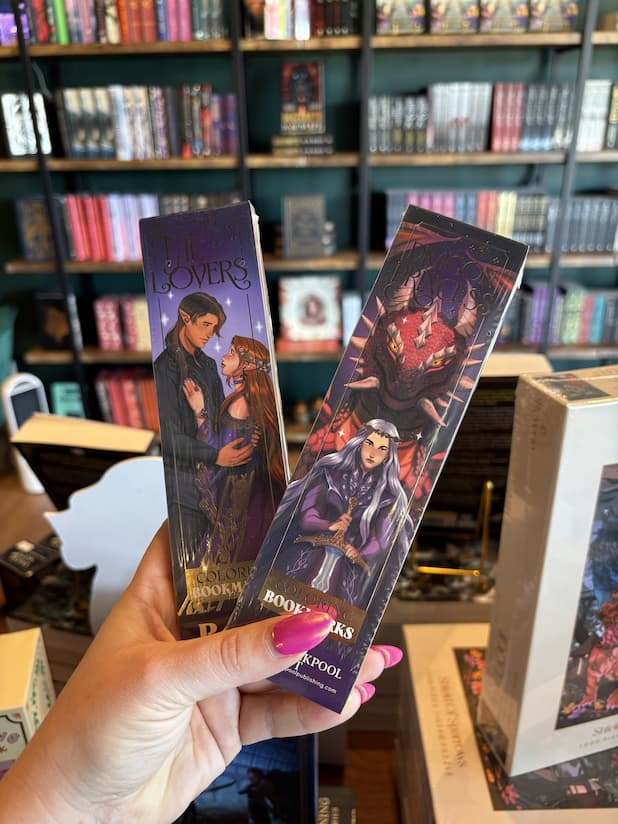 A hand with pink nail polish holds two physical book bookmarks featuring illustrated covers from 'Fae Lovers' and 'Dragon Princess' collections, set against a blurred background of a bookshelves.