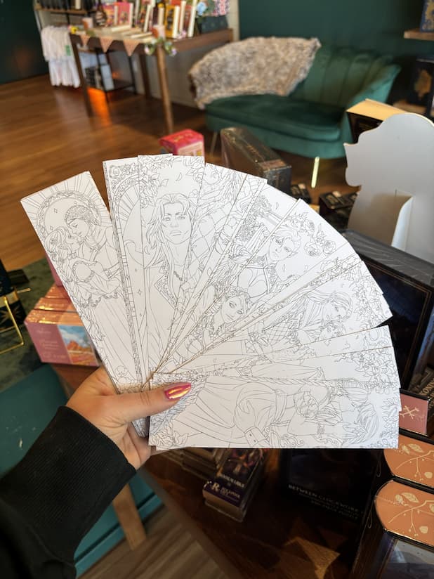 A hand with pink nail polish holds a  fanned-out set bookmarks with black line art illustrations.