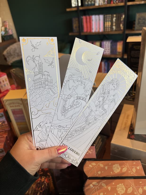 A hand with pink nail polish holds a set of bookmarks with gold foil line art of  illustrations.