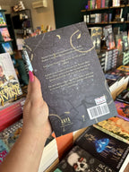 A hand with pink nail polish holding a coy of 'The Shadow Bound Queen: Court of Monsters and Malice' by Eliza Raine with a dark grey background, sharp branches, and yellow text.
