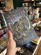 A hand with pink nail polish holding a coy of 'The Shadow Bound Queen: Court of Monsters and Malice' by Eliza Raine with a gold axe, black snakes and gold roses on a dark background.