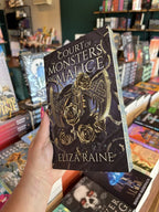 A hand with pink nail polish holding a copy of 'The Shadow Bound Queen: Court of Monsters and Malice' by Eliza Raine with a gold axe, black snakes and gold roses on a dark background with matching black and gold roses on the sprayed edge.