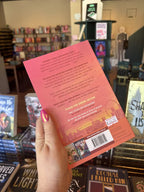 A hand with pink nail polish holding the back of a paperback copy of 'Crash Into You' by Simone Soltani, featuring a sunset background with a city landscape and dark text.