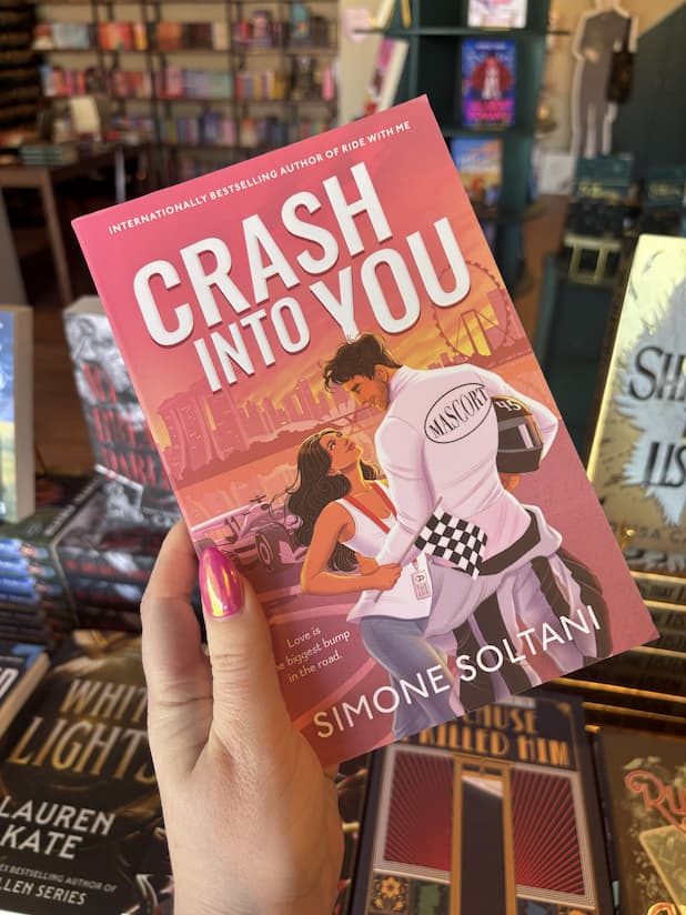 A hand with pink nail polish holding a paperback copy of 'Crash Into You' by Simone Soltani, featuring an illustrated man in a racing uniform embracing a woman by the waist gazing into each other's eyes, in a racing-themed city scene with large yellow title text and the author's name at the bottom.
