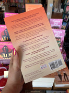A hand holds the back cover of 'Cross Check' by Chelsea Curto, featuring a peach-orange background and black text. The background is a blurred bookstore.