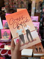 A hand holds the ook cover for 'Cross Check' by Chelsea Curto, featuring an illustrated man and woman on an ice rink with hockey sticks, set against a peach-orange gradient background with the title in large red script. In the background is a blurry bookstore.