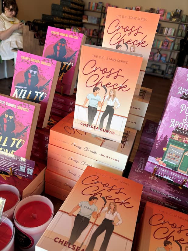 Multiple stacks of 'Cross Check' by Chelsea Curto displayed in a bookstore, featuring an illustrated cover with a man and woman on an ice rink with hockey sticks, set against a peach-orange gradient background with the title in large red script.