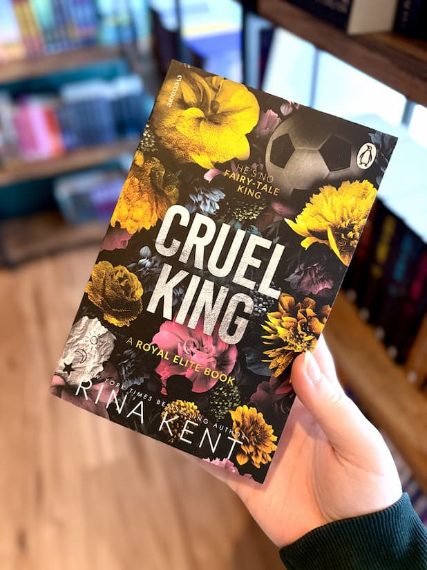 A hand holding a paperback copy of 'Cruel King' by Rina Kent, featuring large textured silver title text over a dark floral background with yellow, pink, purple, and blue flowers, plus a soccer ball graphic.